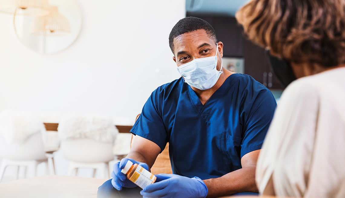 During the coronavirus epidemic, a mid adult male visiting home health nurse discusses medicine side effects with unrecognizable senior woman.  They are both wearing protective masks and the nurse is also wearing protective gloves to prevent the spread of illness.